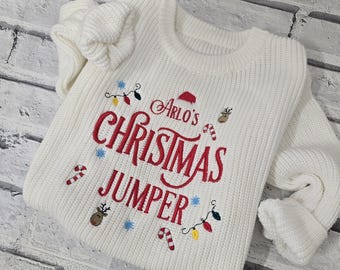 May include: White Christmas sweater with red embroidered text that says "Arlo's Christmas Jumper" and festive designs including candy canes, snowflakes, reindeer, and Christmas lights.