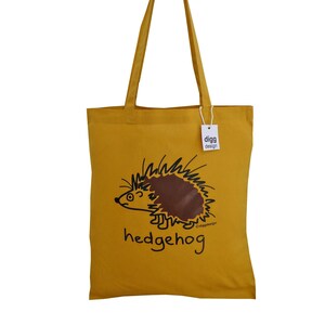 May include: A yellow canvas tote bag with a brown hedgehog illustration and the text "hedgehog" printed on the front. The bag has a white tag with the text "digg design" printed on it.