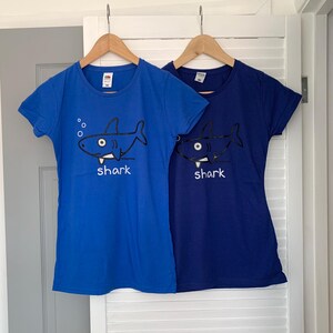 May include: Two blue t-shirts featuring a cartoon shark design. The shark is black and white, with the word "shark" printed underneath. One shirt is a lighter blue, the other a darker blue. Both are hanging on wooden hangers.