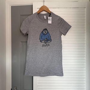 May include: A heather grey short-sleeved t-shirt featuring a cartoon illustration of a blue sloth sitting down. Below the sloth, the word "sloth" is printed in black lowercase letters. A white tag with the letters "dd" is attached to the collar.