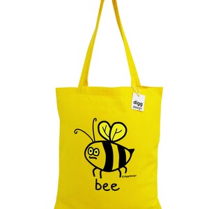 May include: A yellow tote bag with a black and yellow bee illustration and the word "bee" printed on it. The bag has a black tag with the text "digg design" printed on it.