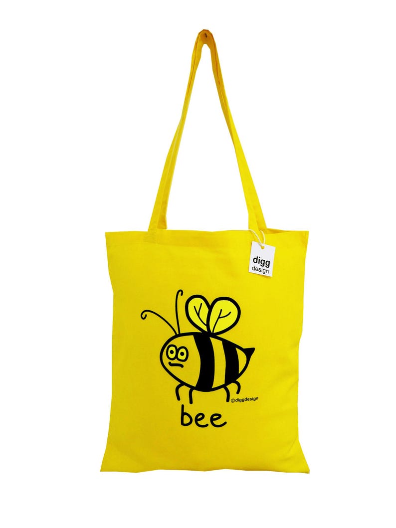 May include: A yellow tote bag with a black and yellow bee illustration and the word "bee" printed on it. The bag has a black tag with the text "digg design" printed on it.
