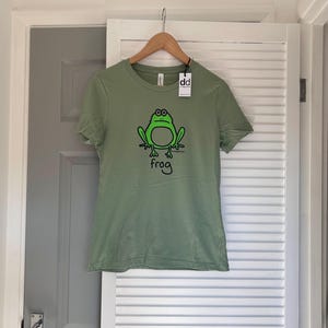 Pu&ograve; includere: T-shirt verde salvia con un'illustrazione di rana dei cartoni animati in nero e verde. La parola "frog" &egrave; scritta sotto la rana. La maglietta &egrave; appesa a una gruccia di legno con un'etichetta "dd".