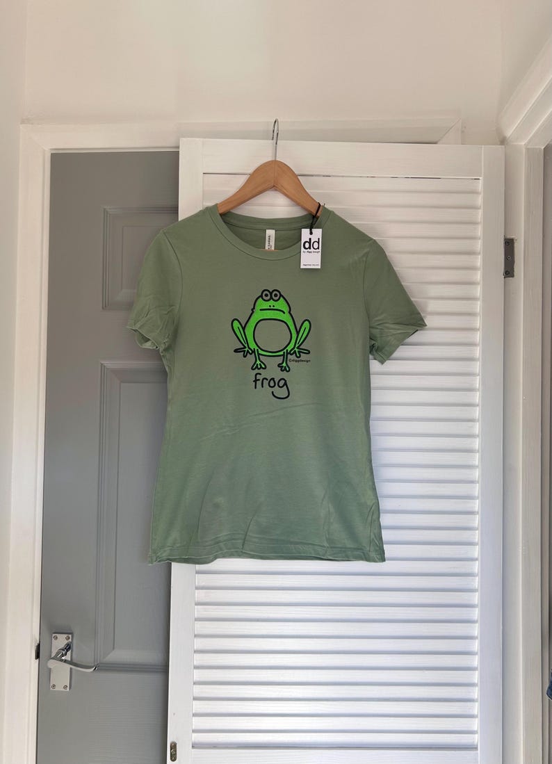 Pu&ograve; includere: T-shirt verde salvia con un'illustrazione di rana dei cartoni animati in nero e verde. La parola "frog" &egrave; scritta sotto la rana. La maglietta &egrave; appesa a una gruccia di legno con un'etichetta "dd".