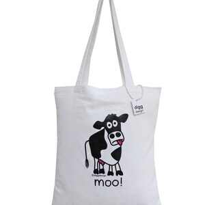 May include: White canvas tote bag with a black and white cartoon cow with a pink tongue sticking out. The cow is printed on the bag with the text "moo!" underneath. The bag has a white handle and a small white tag with the text "digg design" on it.