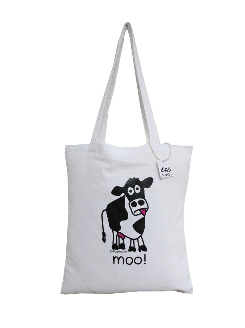 May include: White canvas tote bag with a black and white cartoon cow with a pink tongue sticking out. The cow is printed on the bag with the text "moo!" underneath. The bag has a white handle and a small white tag with the text "digg design" on it.
