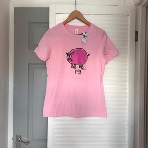 Pu&ograve; includere: T-shirt rosa chiaro con un disegno di maiale dei cartoni animati. Il maiale &egrave; rosa con contorni neri e la parola "pig" &egrave; scritta sotto. La maglietta &egrave; appesa a un appendiabiti di legno.