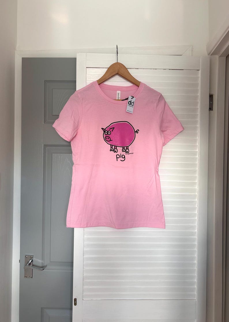 Pu&ograve; includere: T-shirt rosa chiaro con un disegno di maiale dei cartoni animati. Il maiale &egrave; rosa con contorni neri e la parola "pig" &egrave; scritta sotto. La maglietta &egrave; appesa a un appendiabiti di legno.