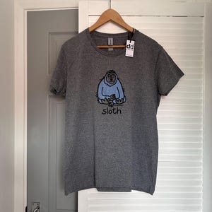 May include: A heather grey short-sleeved t-shirt featuring a graphic of a light blue sloth with a black outline. Below the sloth, the word "sloth" is printed in black lowercase letters. A white tag with the letters "dd" is attached to the collar.