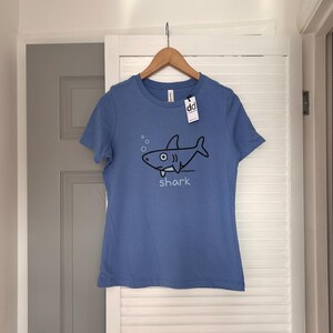 Pu&ograve; includere: Maglietta azzurra con grafica di uno squalo dei cartoni animati. L'illustrazione dello squalo nero include bolle e la parola "shark" sotto. La maglietta &egrave; appesa a un appendiabiti di legno.