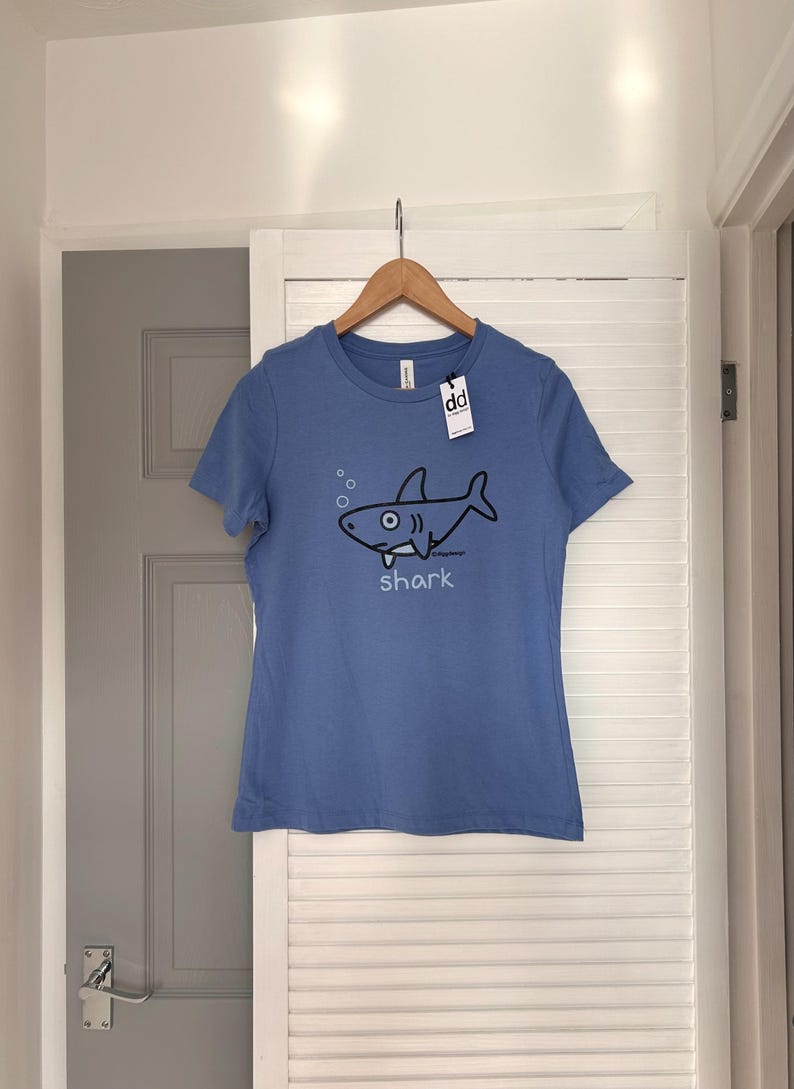 Pu&ograve; includere: Maglietta azzurra con grafica di uno squalo dei cartoni animati. L'illustrazione dello squalo nero include bolle e la parola "shark" sotto. La maglietta &egrave; appesa a un appendiabiti di legno.