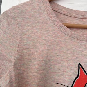Pu&ograve; includere: T-shirt rosa chiaro con girocollo e maniche corte. La maglietta presenta una grafica di gatto rosso con dettagli neri. Il tessuto ha un aspetto m&eacute;lange, con sottili variazioni di colore.