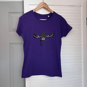 May include: A short-sleeved purple t-shirt featuring a graphic of a black bat hanging upside down with white wing details and large white eyes. Below the bat, the word bat is printed in lowercase black letters on the front of the garment.