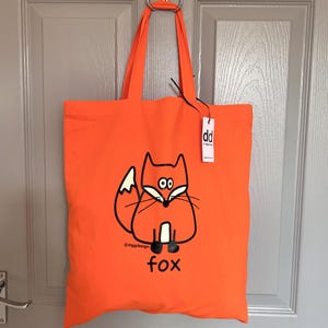 Può includere: Borsa tote arancione brillante con un'illustrazione di volpe dei cartoni animati. La volpe è in bianco e nero, con la parola "fox" sotto. La borsa ha manici lunghi e un'etichetta con "dd".