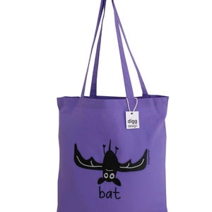 May include: Purple tote bag with a black bat illustration and the word "bat" printed on the front. The bag has a white tag with the text "digg design" on it.