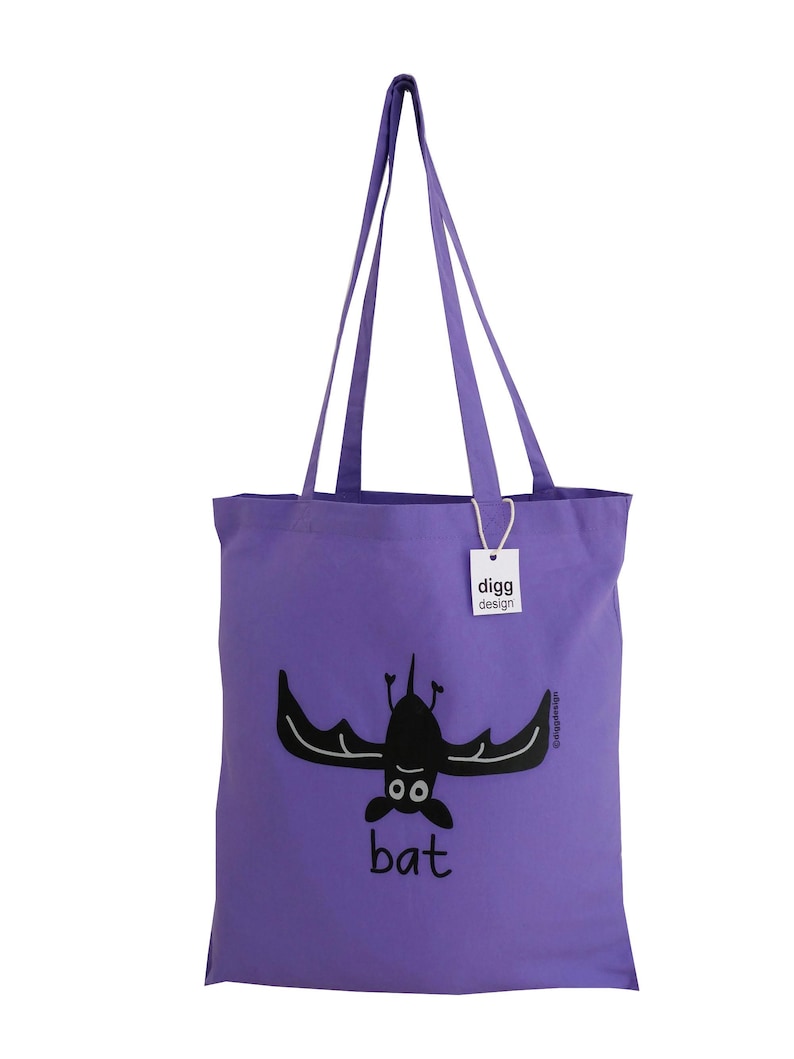 May include: Purple tote bag with a black bat illustration and the word "bat" printed on the front. The bag has a white tag with the text "digg design" on it.