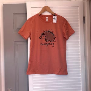 Pu&ograve; includere: T-shirt arancione a maniche corte con un'illustrazione nera di un riccio e la parola "hedgehog". La maglietta &egrave; appesa a una gruccia di legno. L'etichetta dice "dd".