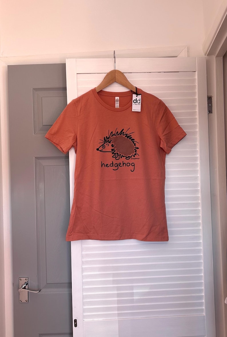 Pu&ograve; includere: T-shirt arancione a maniche corte con un'illustrazione nera di un riccio e la parola "hedgehog". La maglietta &egrave; appesa a una gruccia di legno. L'etichetta dice "dd".