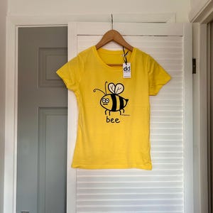 Pu&ograve; includere: Maglietta gialla con un disegno di ape dei cartoni animati in nero e giallo, con la parola "bee" sotto. La maglietta &egrave; appesa a un appendiabiti di legno davanti a una porta bianca.