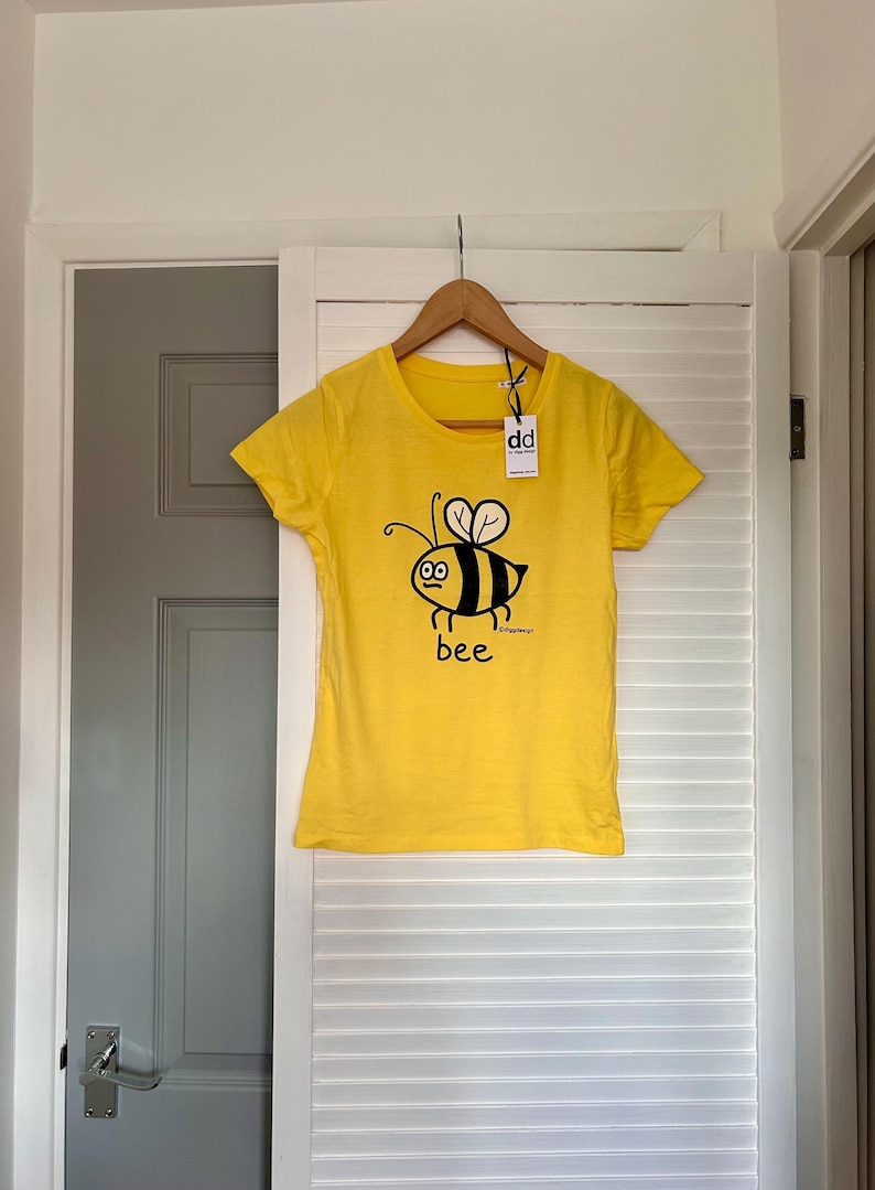 Pu&ograve; includere: Maglietta gialla con un disegno di ape dei cartoni animati in nero e giallo, con la parola "bee" sotto. La maglietta &egrave; appesa a un appendiabiti di legno davanti a una porta bianca.