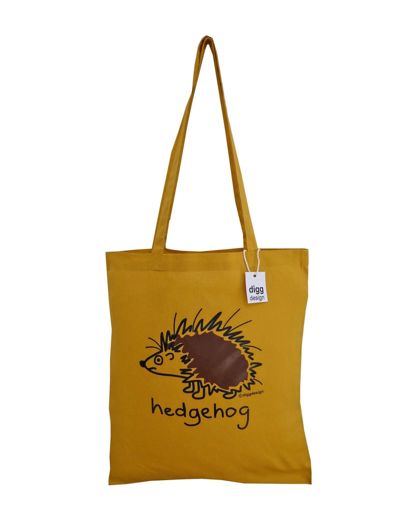 May include: A yellow canvas tote bag with a brown hedgehog illustration and the text "hedgehog" printed on the front. The bag has a white tag with the text "digg design" printed on it.