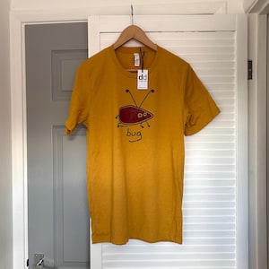 Può includere: T-shirt giallo senape con un disegno di insetto dei cartoni animati. L'insetto è rosso con antenne e occhi neri, e la parola "bug" è scritta sotto. La maglietta è appesa a un appendiabiti di legno.