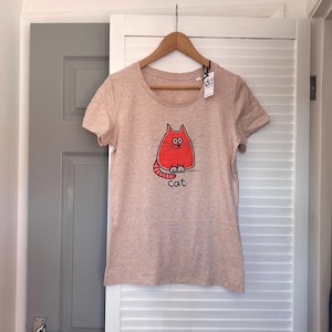 Pu&ograve; includere: T-shirt rosa chiaro con scollo tondo, appesa a una gruccia di legno. La maglietta presenta un'illustrazione di un gatto dei cartoni animati rosso con la parola "cat" sotto. Un'etichetta &egrave; attaccata alla gruccia.
