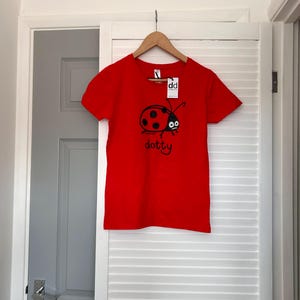 Pu&ograve; includere: Maglietta rosso vivo con grafica di coccinella nera e la scritta "dotty". La maglietta &egrave; appesa a una gruccia di legno. Un'etichetta bianca con "dd" &egrave; attaccata. La maglietta &egrave; appesa davanti a una porta a lamelle bianche.