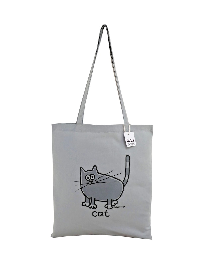 May include: A gray canvas tote bag with a cartoon cat design. The cat is gray with black eyes and a black outline. The bag has a gray handle and the text "digg design" is printed on a white tag attached to the bag. The text "cat" is printed below the cat.