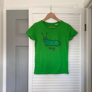 Pu&ograve; includere: Una maglietta verde brillante con un disegno di lumaca dei cartoni animati. L'illustrazione &egrave; una lumaca verde con antenne nere e la parola "slug" sotto. La maglietta &egrave; appesa a una gruccia di legno.