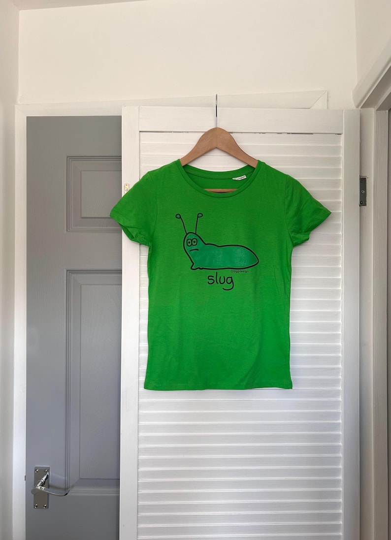 Pu&ograve; includere: Una maglietta verde brillante con un disegno di lumaca dei cartoni animati. L'illustrazione &egrave; una lumaca verde con antenne nere e la parola "slug" sotto. La maglietta &egrave; appesa a una gruccia di legno.