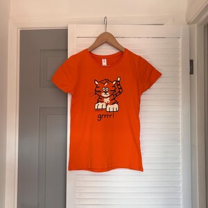 Pu&ograve; includere: T-shirt arancione con un'illustrazione di una tigre dei cartoni animati. La tigre &egrave; arancione e marrone con una zampa alzata e la parola "grrrr!" sotto. La maglietta &egrave; appesa a una gruccia di legno.
