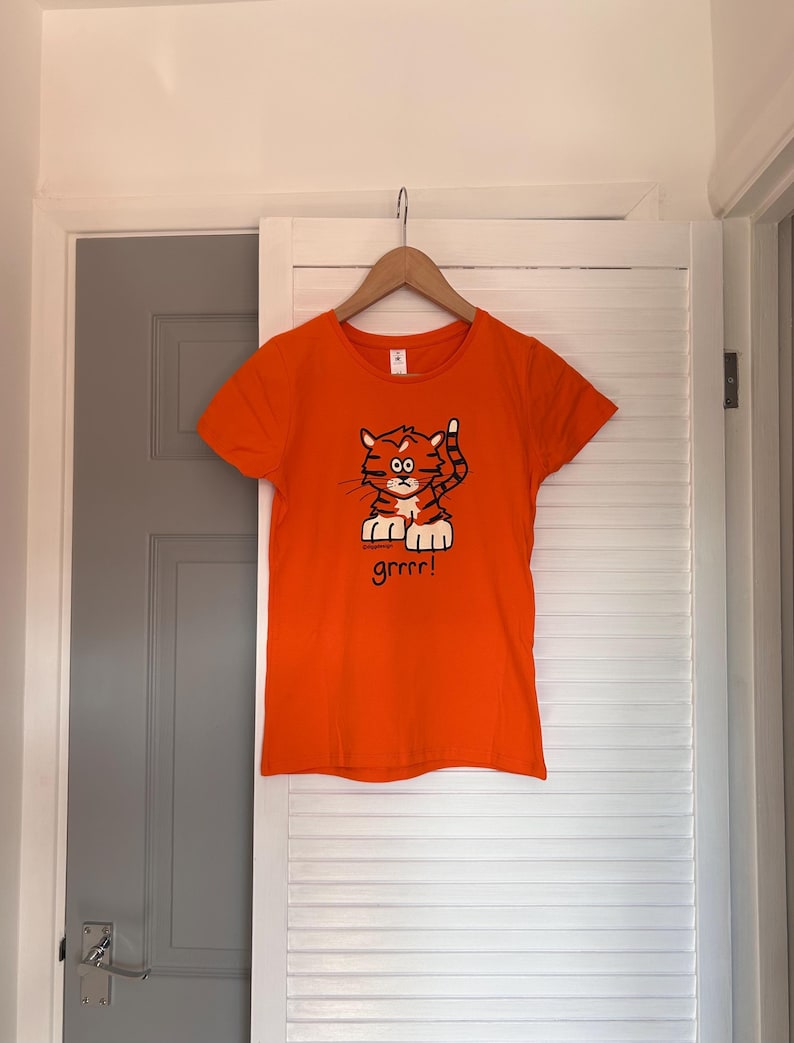 Pu&ograve; includere: T-shirt arancione con un'illustrazione di una tigre dei cartoni animati. La tigre &egrave; arancione e marrone con una zampa alzata e la parola "grrrr!" sotto. La maglietta &egrave; appesa a una gruccia di legno.