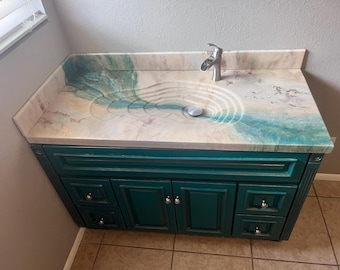 May include: A bathroom vanity cabinet in a deep teal finish with silver-toned knobs, featuring a countertop with a marbled beige and white surface and a recessed, ripple-patterned sink basin accented by a flowing teal resin design and a silver faucet.