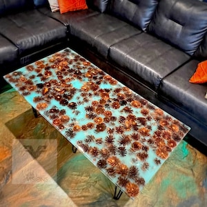 May include: A turquoise coffee table with a resin top featuring a design of brown pine cones. The table has black hairpin legs and is sitting on a colorful rug.