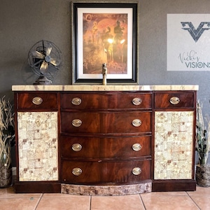 May include: A vintage mahogany sideboard with a marble top and four drawers. The sideboard has been refinished with a decorative paper design on the doors. The sideboard is in front of a gray wall with a framed print and a decorative fan.
