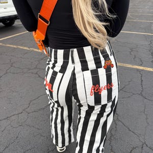 May include: Black and white vertically striped pants featuring two back pockets. The left pocket displays the red text PHL, while the right pocket features the red text flyers and a small orange mascot graphic wearing a black hat.