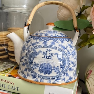 May include: A white ceramic teapot featuring a blue floral pattern and a central oval design with Chinese characters and a decorative incense burner illustration. The teapot has a curved spout, a lid with a small knob, and a tan woven rattan handle.