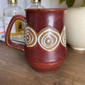 May include: A cylindrical ceramic mug with a deep reddish-brown glaze featuring a horizontal band of repeating white and tan circular swirl patterns. The mug has a sturdy, curved handle on the left side and a smooth, slightly flared rim at the top.