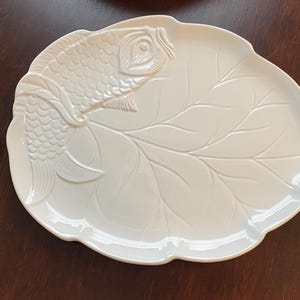 May include: A white ceramic serving platter shaped like a large leaf, featuring a raised, embossed koi fish design on the left side. The surface of the platter displays detailed vein patterns of a leaf, with a smooth, glossy finish throughout.