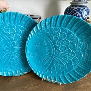 Puede incluir: Dos platos redondos de cerámica de color turquesa brillante con un diseño de pez en relieve. Los platos muestran patrones detallados de escamas, aletas y un ojo prominente, con un borde festoneado que imita la forma de una cola de pez. El acabado brillante refleja la luz sobre la superficie texturizada de los platos, que se exhiben en posición vertical sobre pequeños soportes de metal dorado.