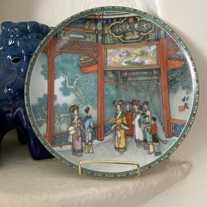 May include: Decorative plate featuring a detailed scene of figures in traditional Chinese attire within a palace setting. The plate has a colorful border and is displayed on a gold-colored stand. The scene includes text in Chinese characters.