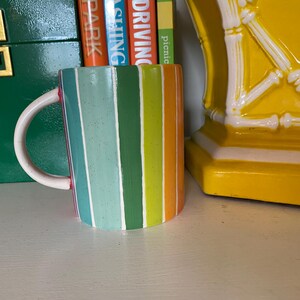 Puede incluir: Una taza de cer&aacute;mica colorida con rayas verticales en tonos azules, verdes, naranjas y amarillos. La taza tiene un asa blanca y un borde rojo. La taza est&aacute; sobre una superficie blanca.