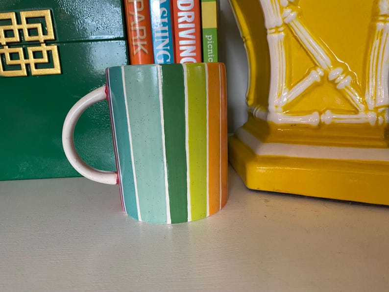 Puede incluir: Una taza de cer&aacute;mica colorida con rayas verticales en tonos azules, verdes, naranjas y amarillos. La taza tiene un asa blanca y un borde rojo. La taza est&aacute; sobre una superficie blanca.