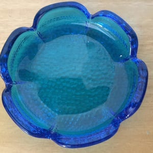 Può includere: Un posacenere di vetro blu con un bordo a forma di fiore. Il posacenere ha una superficie testurizzata e una ciotola profonda.