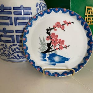 May include: A white decorative plate with a gold rim, featuring a floral design of pink blossoms on a black branch. The plate has a blue and pink patterned border. It is displayed on a gold metal stand.