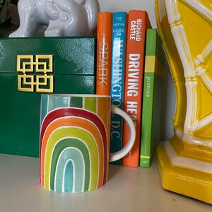 Puede incluir: Una taza de cer&aacute;mica con un dise&ntilde;o de arco&iacute;ris en tonos rojos, naranjas, amarillos, verdes y azules. La taza tiene un asa blanca y est&aacute; colocada cerca de libros y objetos decorativos.