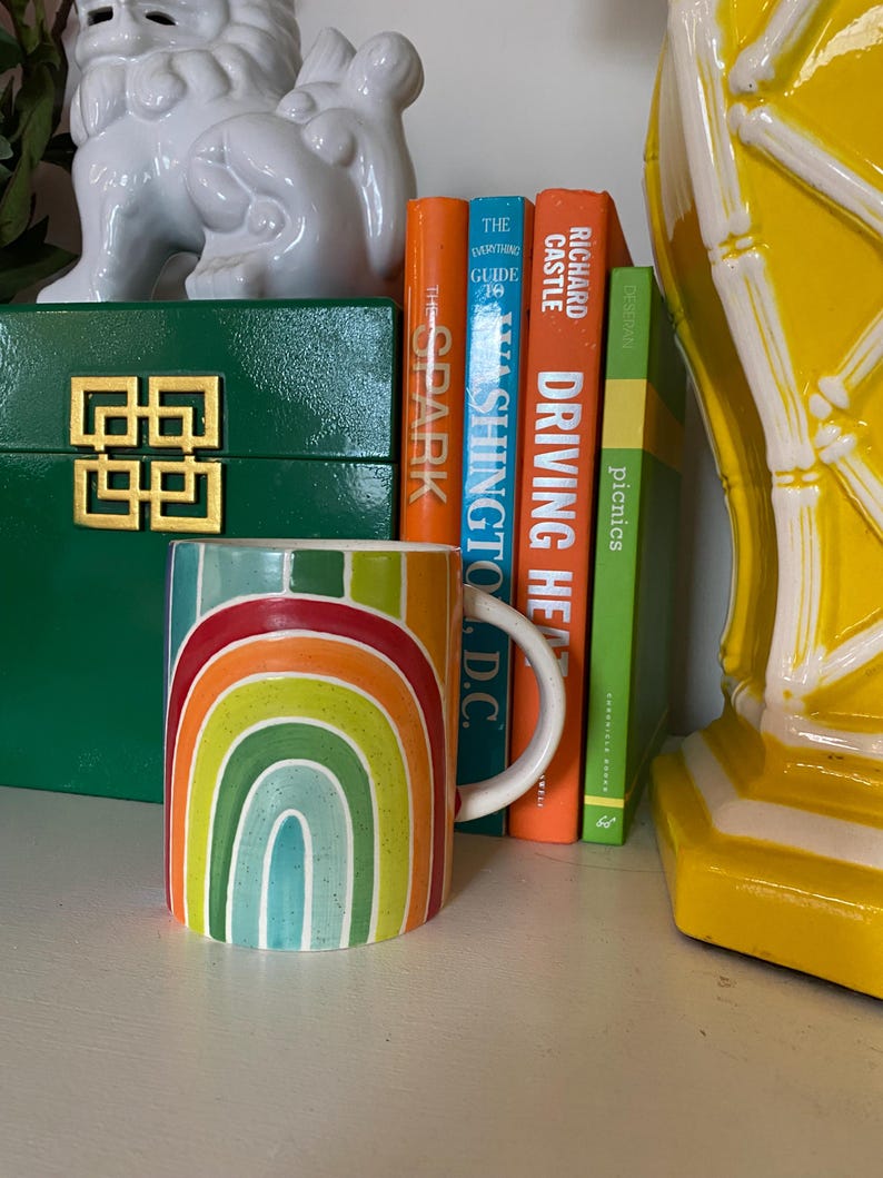 Puede incluir: Una taza de cer&aacute;mica con un dise&ntilde;o de arco&iacute;ris en tonos rojos, naranjas, amarillos, verdes y azules. La taza tiene un asa blanca y est&aacute; colocada cerca de libros y objetos decorativos.