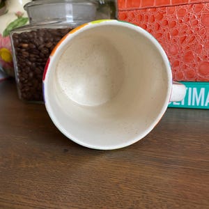 Puede incluir: Una taza de cer&aacute;mica moteada con un borde de color arco&iacute;ris. La taza es blanca con un patr&oacute;n moteado y un asa peque&ntilde;a. La taza est&aacute; sobre una superficie de madera marr&oacute;n.