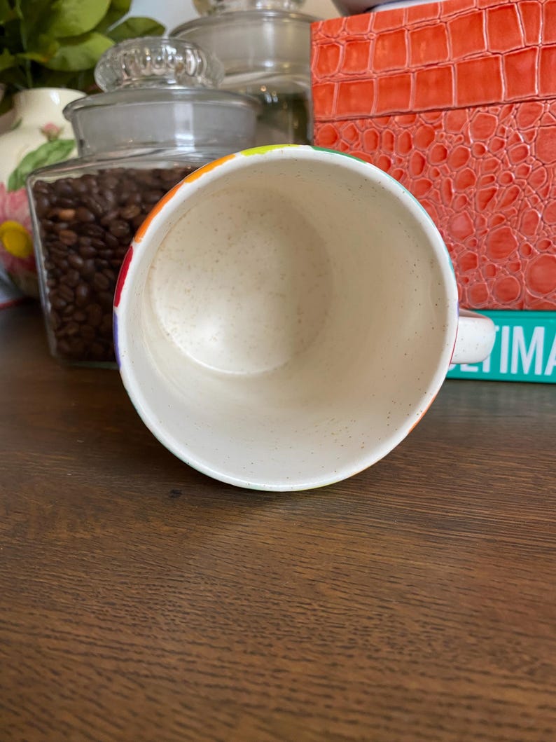 Puede incluir: Una taza de cer&aacute;mica moteada con un borde de color arco&iacute;ris. La taza es blanca con un patr&oacute;n moteado y un asa peque&ntilde;a. La taza est&aacute; sobre una superficie de madera marr&oacute;n.